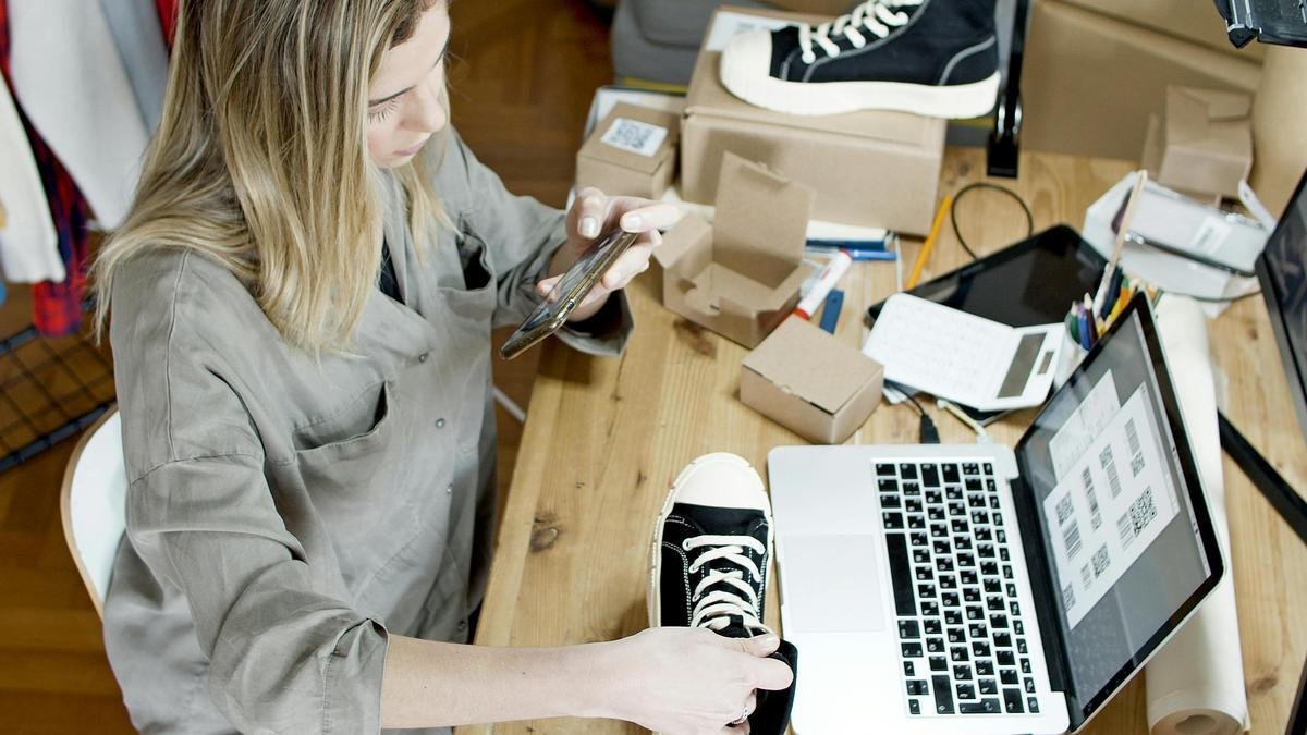 Mulher tirando foto de um tênis para venda online em uma bancada, usando smartphone e laptop, com caixas e materiais de embalagem ao redor